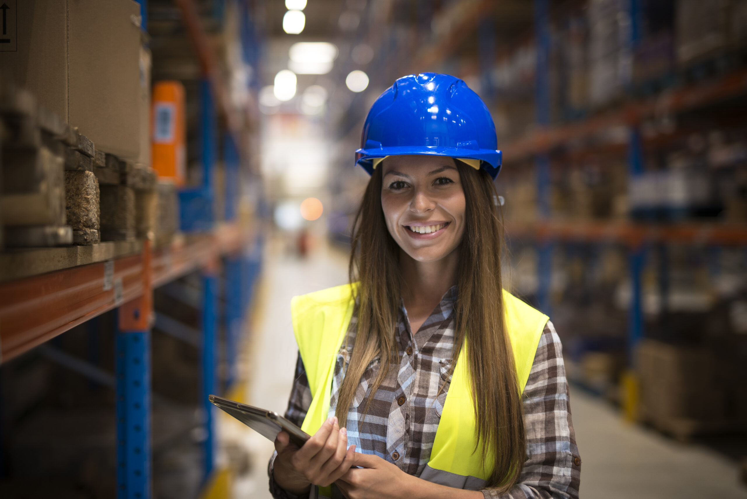 Portrait of smiling woman in protective uniform with hardhat holding tablet in warehouse center.