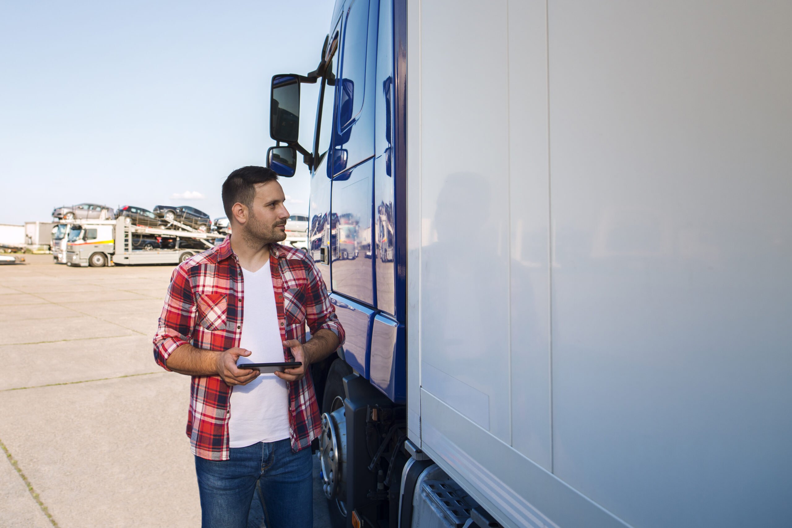 Truck driver in casual clothes standing by his truck with tablet and looking at tarpaulin.